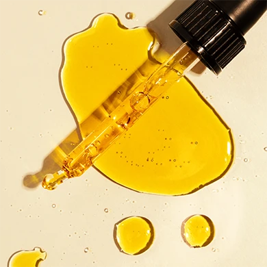 Golden yellow liquid droplets on a light beige surface with a black dropper pipette partially visible at the top right.